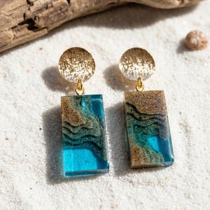 Puede incluir: Pendientes dorados con una parte superior circular y un colgante rectangular. El colgante presenta un diseño azul y marrón, que se asemeja a un paisaje en miniatura. Los pendientes están sobre un fondo de arena blanca.