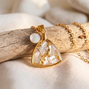 Puede incluir: Un collar dorado con un colgante triangular lleno de trozos iridiscentes de concha y copos de oro. El colgante está enmarcado en oro y cuelga de una delicada cadena. Un pequeño dije blanco y redondo cuelga cerca del colgante.