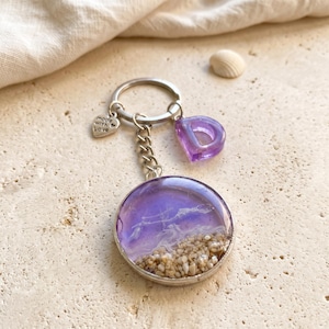 Pode incluir: Um porta-chaves circular com uma cena de oceano em resina roxa com areia na parte inferior, preso a um anel de metal prateado. Inclui um pequeno pendente com a letra D roxa e um pendente prateado em forma de coração gravado made with Love.