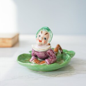 Puede incluir: Figura de cerámica vintage de un elfo sonriente con un atuendo morado y blanco, descansando sobre un plato verde con forma de hoja. El elfo tiene un sombrero verde y está acostado. Un jarrón azul con bayas naranjas en el fondo.