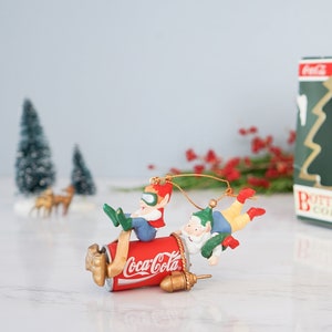 Op de afbeelding: Een vintage Coca-Cola kerstversiering met twee elven die op een Coca-Cola-blik rijden. De versiering is goud, rood en groen en wordt geleverd in een doos met de tekst "Bottle Works Collection".