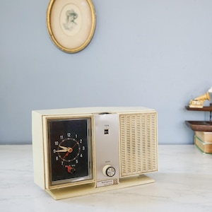 Könnte beinhalten: Vintage cremefarbenes Radiowecker mit schwarzem Zifferblatt und roten Akzenten. Die Uhr hat ein Retro-Design mit Lautsprecher und Einstellrad. Ein dekorativer Artikel für ein Schlaf- oder Wohnzimmer.