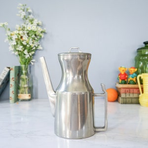 以下が含まれることがあります： 蓋と取っ手が付いた銀色の金属製のティーポット。ティーポットは白い大理石の表面の上に置かれています。