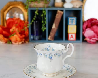 May include: A white teacup and saucer set with a delicate blue floral pattern and gold trim. The teacup has a curved handle and sits on a matching saucer. The set is displayed on a marble surface.