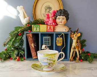 May include: A vintage teacup and saucer with a floral design sits on a white surface.  A wooden shelf with a book, a birdcage, and a doll are in the background.  The shelf is decorated with red berries and green pine branches.  The blocks on the shelf spell out "TEA".