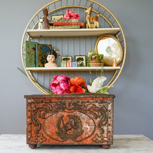 Può includere: Un ripiano circolare in metallo dorato a tre livelli. Il ripiano è decorato con fiori, libri, una bambola e una piccola statuetta di cervo. Il ripiano è posto sopra una scatola di metallo vintage rossa con dettagli decorativi.