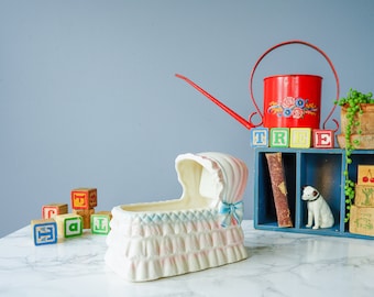 May include: A white ceramic baby cradle with blue and pink trim sits on a white marble surface.  A red watering can with a floral design and a wooden shelf with blocks spelling "TREE" are in the background.