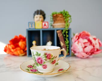 Puede incluir: Una taza de té y un platillo blancos con un patrón floral de rosas rosadas y hojas verdes. La taza de té tiene un borde dorado y el platillo tiene una banda dorada alrededor del borde. La taza de té y el platillo están sobre una superficie de mármol blanco.