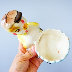 May include: A ceramic chick figurine with a yellow body, brown hat, and blue and white skirt. The chick is holding a white egg with a blue rim.
