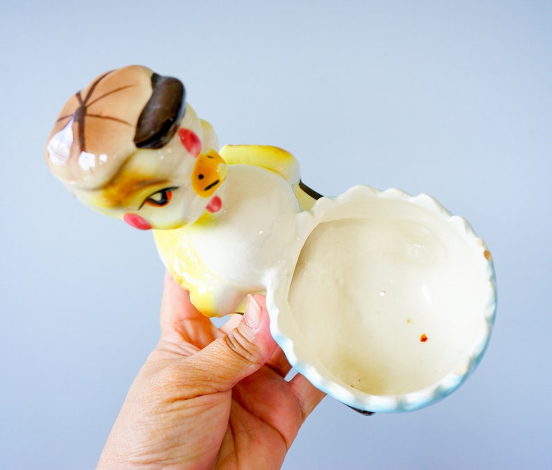 May include: A ceramic chick figurine with a yellow body, brown hat, and blue and white skirt. The chick is holding a white egg with a blue rim.