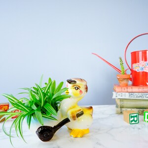 May include: A yellow ceramic planter shaped like a duck pulling a wagon, with a green spider plant growing inside. The planter is on a white marble surface with red and green alphabet blocks and a red watering can with a cactus in a terracotta pot on top of a stack of books.