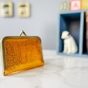 Puede incluir: Un bolso de mano dorado con un estampado floral decorativo. El bolso tiene un cierre de metal y un acabado liso y brillante.