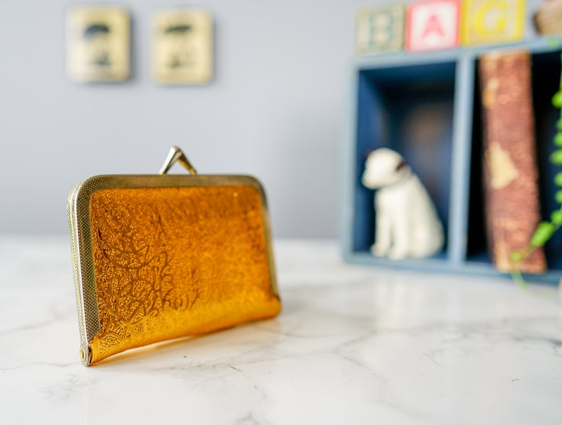 Puede incluir: Un bolso de mano dorado con un estampado floral decorativo. El bolso tiene un cierre de metal y un acabado liso y brillante.