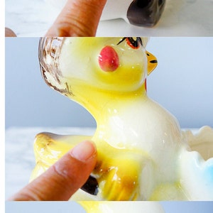 May include: A yellow ceramic chick pushing a blue and white egg-shaped planter. The chick has a brown beak, black eyes, and a pink blush on its cheeks. The planter has a scalloped edge.