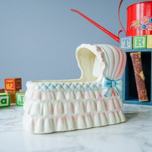 May include: A white ceramic baby crib planter with pink and blue details. The planter has a ruffled edge and a blue ribbon bow. The planter is sitting on a white marble surface.