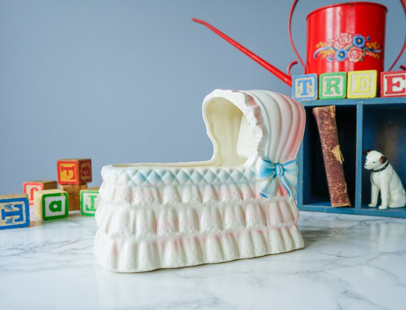 May include: A white ceramic baby crib planter with pink and blue details. The planter has a ruffled edge and a blue ribbon bow. The planter is sitting on a white marble surface.