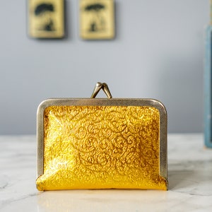Puede incluir: Una cartera de monedas dorada con un patr&oacute;n decorativo en relieve. La cartera tiene un marco de metal plateado y un cierre de presi&oacute;n.