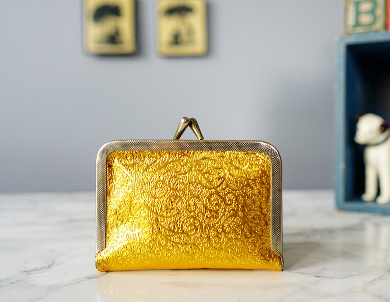 Puede incluir: Una cartera de monedas dorada con un patr&oacute;n decorativo en relieve. La cartera tiene un marco de metal plateado y un cierre de presi&oacute;n.