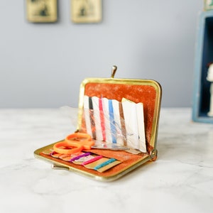 Puede incluir: Un kit de costura de metal dorado con un interior de terciopelo marr&oacute;n. El kit est&aacute; abierto y contiene varios art&iacute;culos de costura, incluidas agujas, hilo y un peque&ntilde;o par de tijeras naranjas.