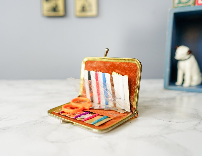 Puede incluir: Un kit de costura de metal dorado con un interior de terciopelo marr&oacute;n. El kit est&aacute; abierto y contiene varios art&iacute;culos de costura, incluidas agujas, hilo y un peque&ntilde;o par de tijeras naranjas.