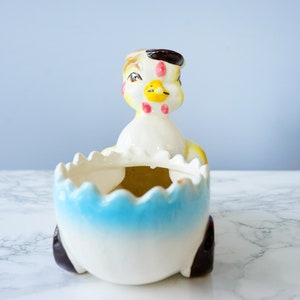 May include: A ceramic chick planter with a blue and white egg-shaped base. The chick has a yellow beak, black eyes, and pink cheeks.