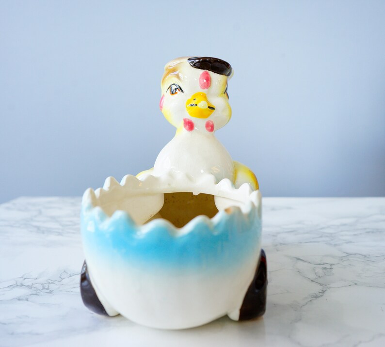 May include: A ceramic chick planter with a blue and white egg-shaped base. The chick has a yellow beak, black eyes, and pink cheeks.