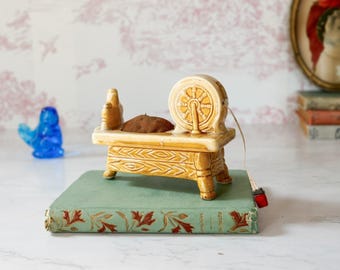 May include: A ceramic spinning wheel figurine in a glossy tan glaze with a brown textured cushion on the base. The piece features a detailed wheel and carved leg accents, resting on a light green hardcover book with decorative leaf patterns.