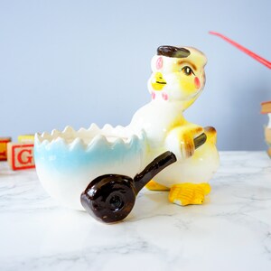 May include: A ceramic chick figurine pushing a blue and white egg-shaped planter. The chick is yellow with brown eyes, a red beak, and pink cheeks. The planter has a brown wheel.