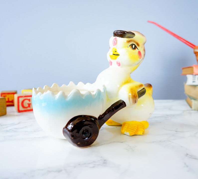 May include: A ceramic chick figurine pushing a blue and white egg-shaped planter. The chick is yellow with brown eyes, a red beak, and pink cheeks. The planter has a brown wheel.
