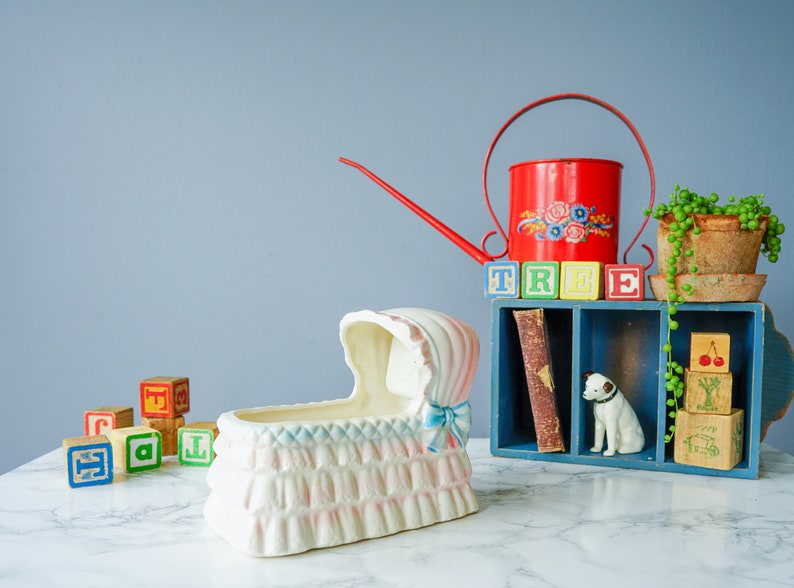 May include: A white ceramic baby cradle with blue and pink trim sits on a white marble surface.  A red watering can with a floral design and a wooden shelf with blocks spelling "TREE" are in the background.