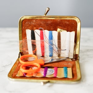 Puede incluir: Un kit de costura vintage de color dorado con un exterior de imitaci&oacute;n de cuero marr&oacute;n. El kit est&aacute; abierto y contiene una variedad de art&iacute;culos de costura, incluyendo tijeras, hilo y botones.