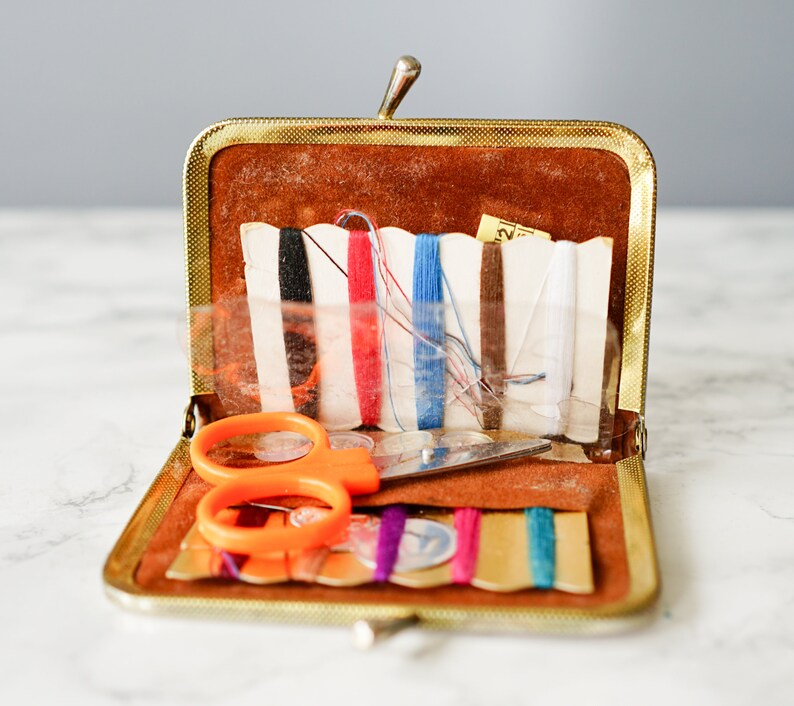 Puede incluir: Un kit de costura vintage de color dorado con un exterior de imitaci&oacute;n de cuero marr&oacute;n. El kit est&aacute; abierto y contiene una variedad de art&iacute;culos de costura, incluyendo tijeras, hilo y botones.