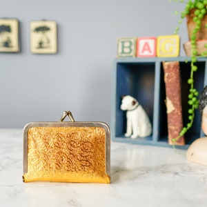 Puede incluir: Una cartera de monedas dorada con una superficie texturizada y un cierre plateado.