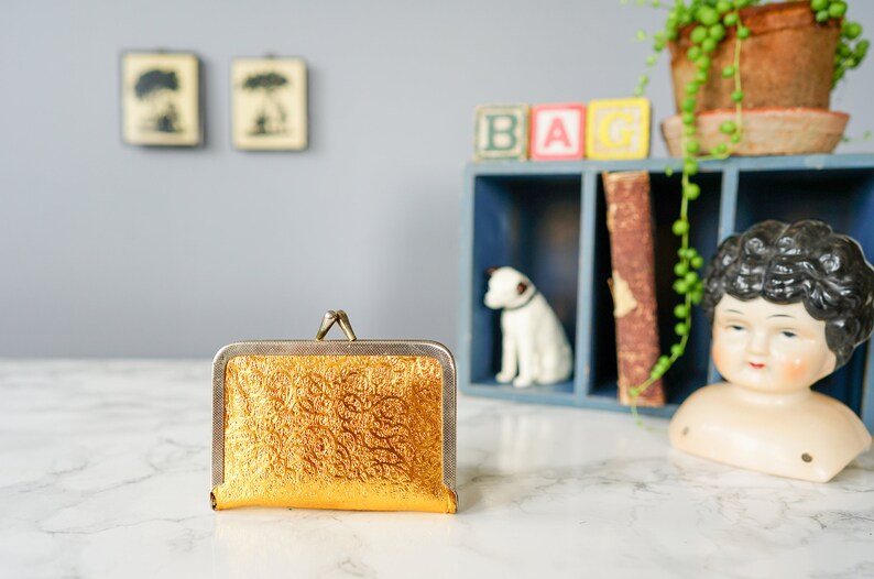 Puede incluir: Una cartera de monedas dorada con una superficie texturizada y un cierre plateado.