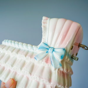 May include: A white ceramic baby carriage with pink and blue details. The carriage has a blue bow and a white ruffle trim. The carriage is a vintage style music box.