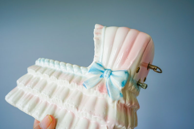 May include: A white ceramic baby carriage with pink and blue details. The carriage has a blue bow and a white ruffle trim. The carriage is a vintage style music box.