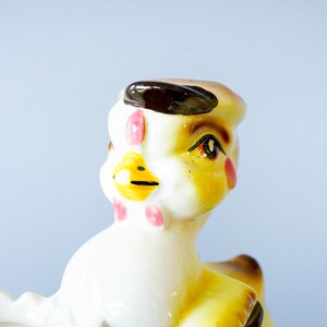 May include: A ceramic chick figurine with a brown hat, yellow body, white face, and pink cheeks. The chick has a yellow beak and brown eyes.