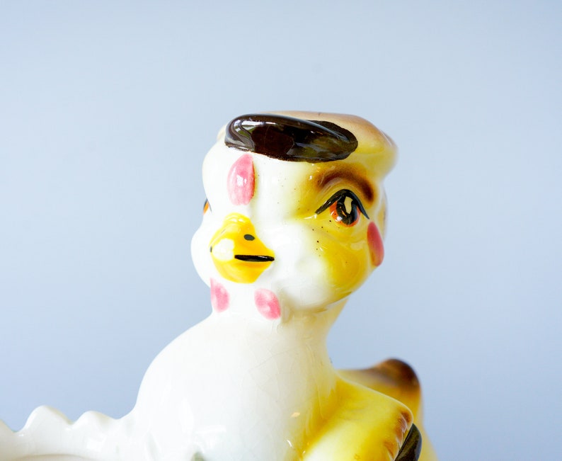 May include: A ceramic chick figurine with a brown hat, yellow body, white face, and pink cheeks. The chick has a yellow beak and brown eyes.