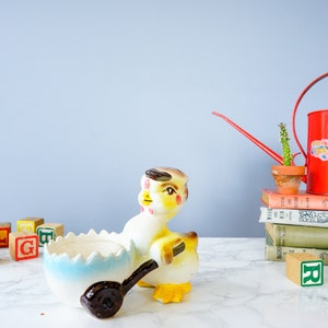 May include: A yellow ceramic chick figurine holding a blue and white egg shaped dish. The chick has brown eyes and a brown beak. The chick is sitting on a white marble surface with colourful wooden blocks and a red watering can with a cactus on a stack of books.