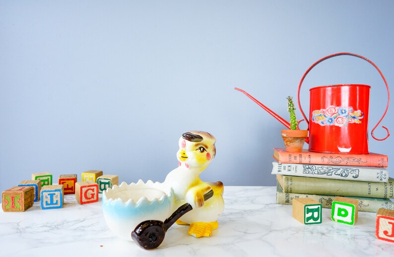 May include: A yellow ceramic chick figurine holding a blue and white egg shaped dish. The chick has brown eyes and a brown beak. The chick is sitting on a white marble surface with colourful wooden blocks and a red watering can with a cactus on a stack of books.