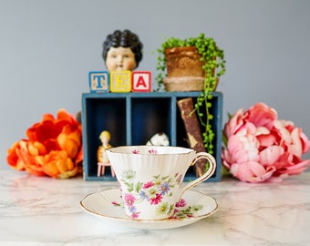 Puede incluir: Una taza de té y un platillo blancos con un patrón floral. La taza de té tiene un borde festoneado y un asa delicada. El platillo tiene un patrón floral a juego y un borde dorado.