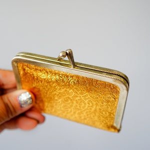 Puede incluir: Una cartera de monedas de color dorado con un cierre plateado y un intrincado patr&oacute;n en relieve.