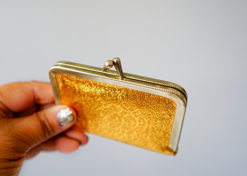 Puede incluir: Una cartera de monedas de color dorado con un cierre plateado y un intrincado patr&oacute;n en relieve.