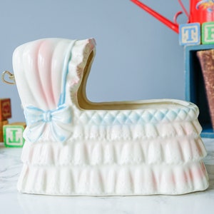 May include: White ceramic baby crib planter with pink and blue trim. The planter has a ruffled edge and a bow on the front.