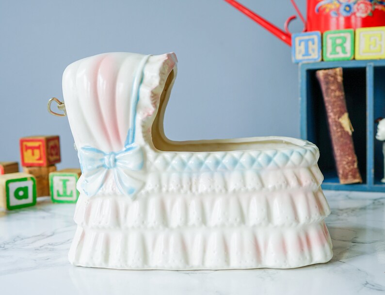 May include: White ceramic baby crib planter with pink and blue trim. The planter has a ruffled edge and a bow on the front.