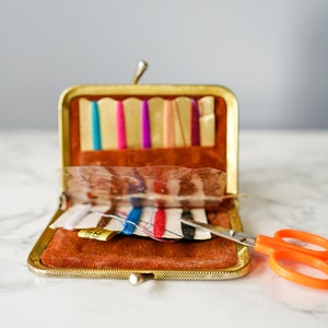 Puede incluir: Un kit de costura de metal dorado con un interior de terciopelo marr&oacute;n. El kit est&aacute; abierto y contiene carretes de hilo, agujas y un par de tijeras naranjas.