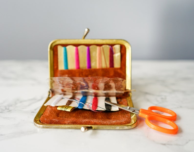 Puede incluir: Un kit de costura de metal dorado con un interior de terciopelo marr&oacute;n. El kit est&aacute; abierto y contiene carretes de hilo, agujas y un par de tijeras naranjas.