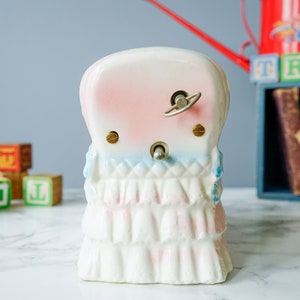 May include: A white porcelain wind-up music box shaped like a girl wearing a pink and blue dress. The music box has a key on the back to wind it up.