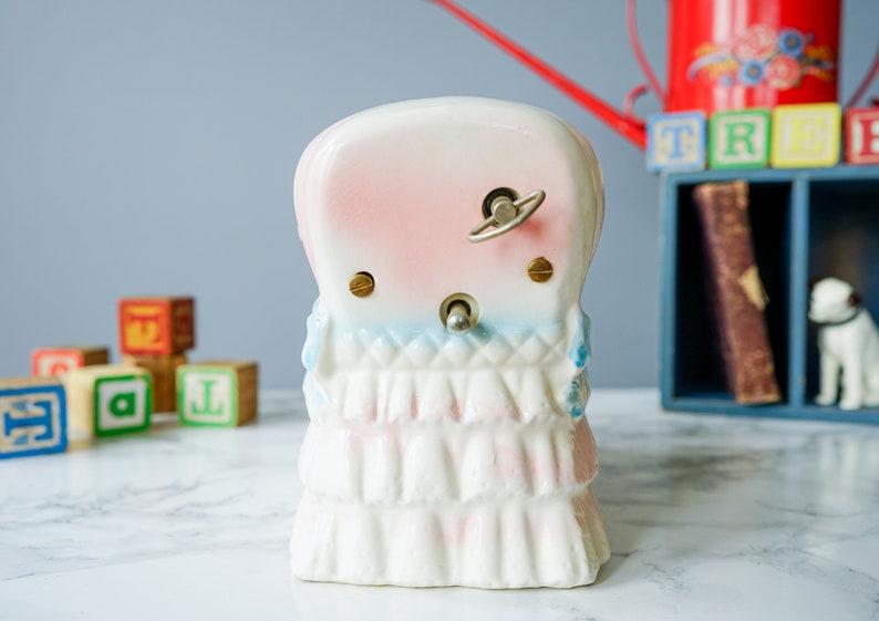 May include: A white porcelain wind-up music box shaped like a girl wearing a pink and blue dress. The music box has a key on the back to wind it up.