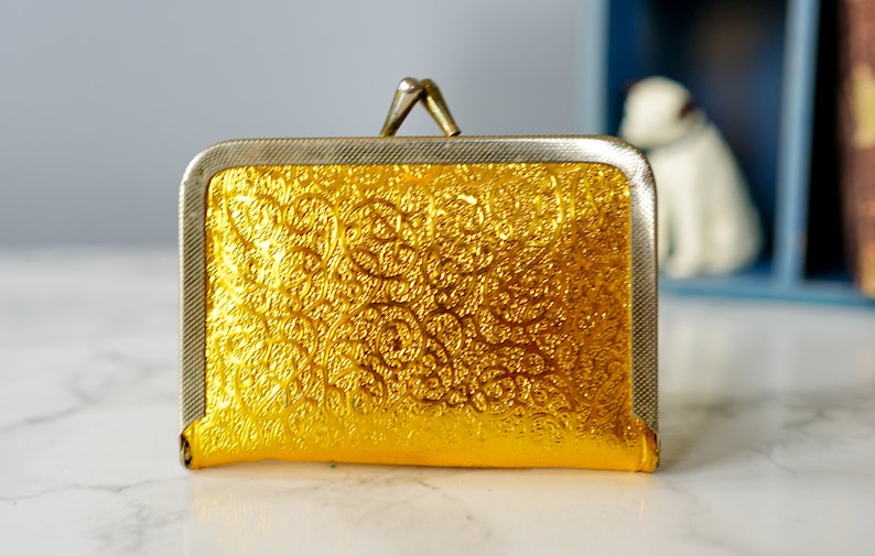 Puede incluir: Una peque&ntilde;a cartera de monedas de oro con un marco de plata y un dise&ntilde;o en relieve floral.